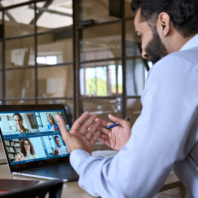 Indian happy smiling businessman ceo having working videocall on planning finance with multiracial team colleagues using laptop pc in modern corporation office. Over shoulder view.